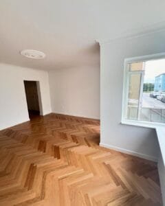 Tom stue med sildebensparketgulv og åbent vindue.